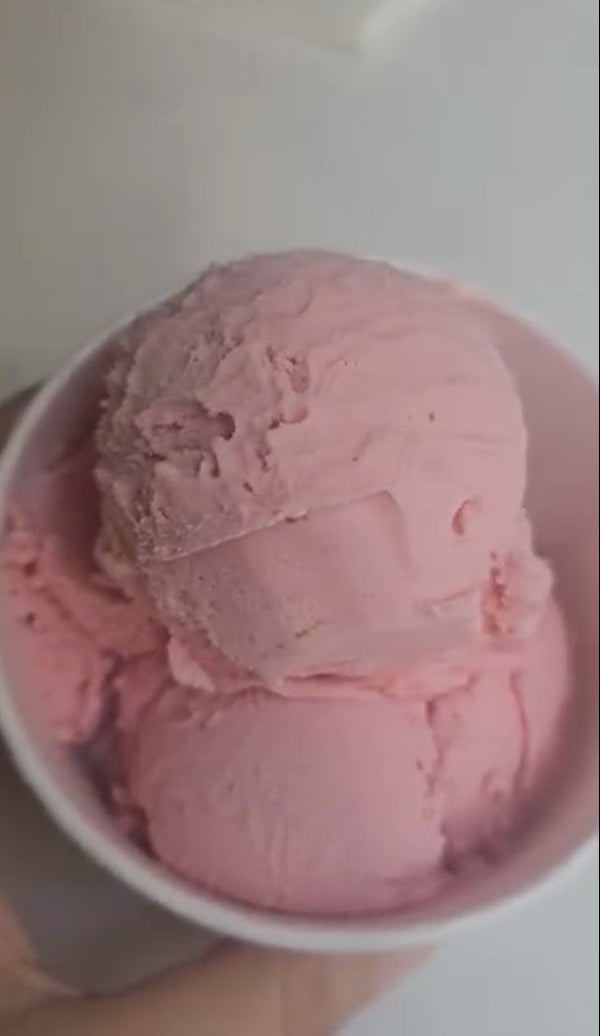A close-up of pink ice cream scoops in a white paper cup held by a hand against a plain light background.